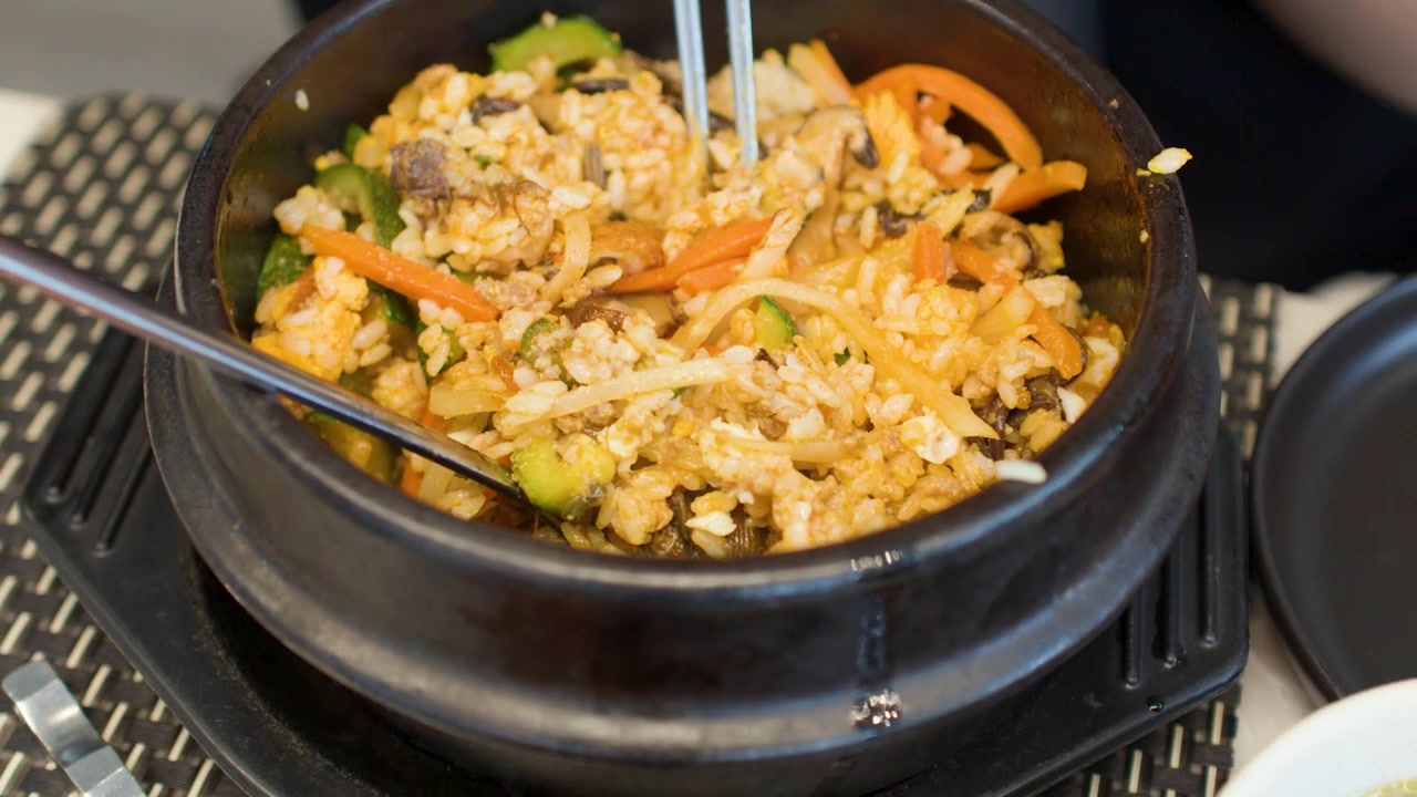 Bidikan close-up pencampuran bibimbap dalam mangkuk batu diikuti oleh berbagai lauk pauk