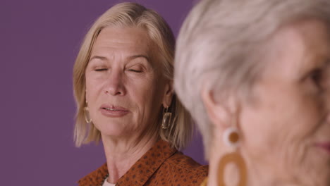 close up view of blonde mature woman with brown shirt posing with blurred senior woman with short hair on purple background