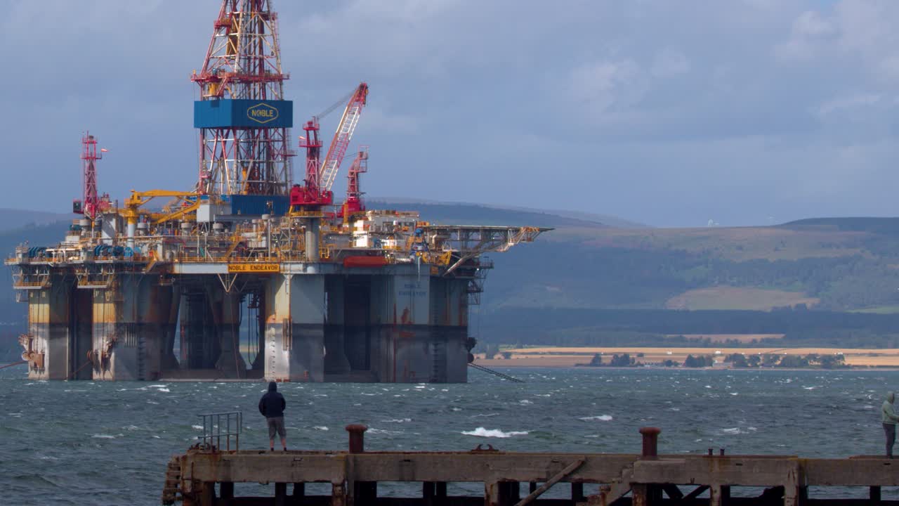 En ensom fisker står på en mole og kigger mod en stor offshore olieplatform i dagslys
