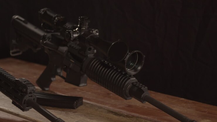 Dolly out of AR 15 rifle with scope, revealing barrel of other rifle on wooden table