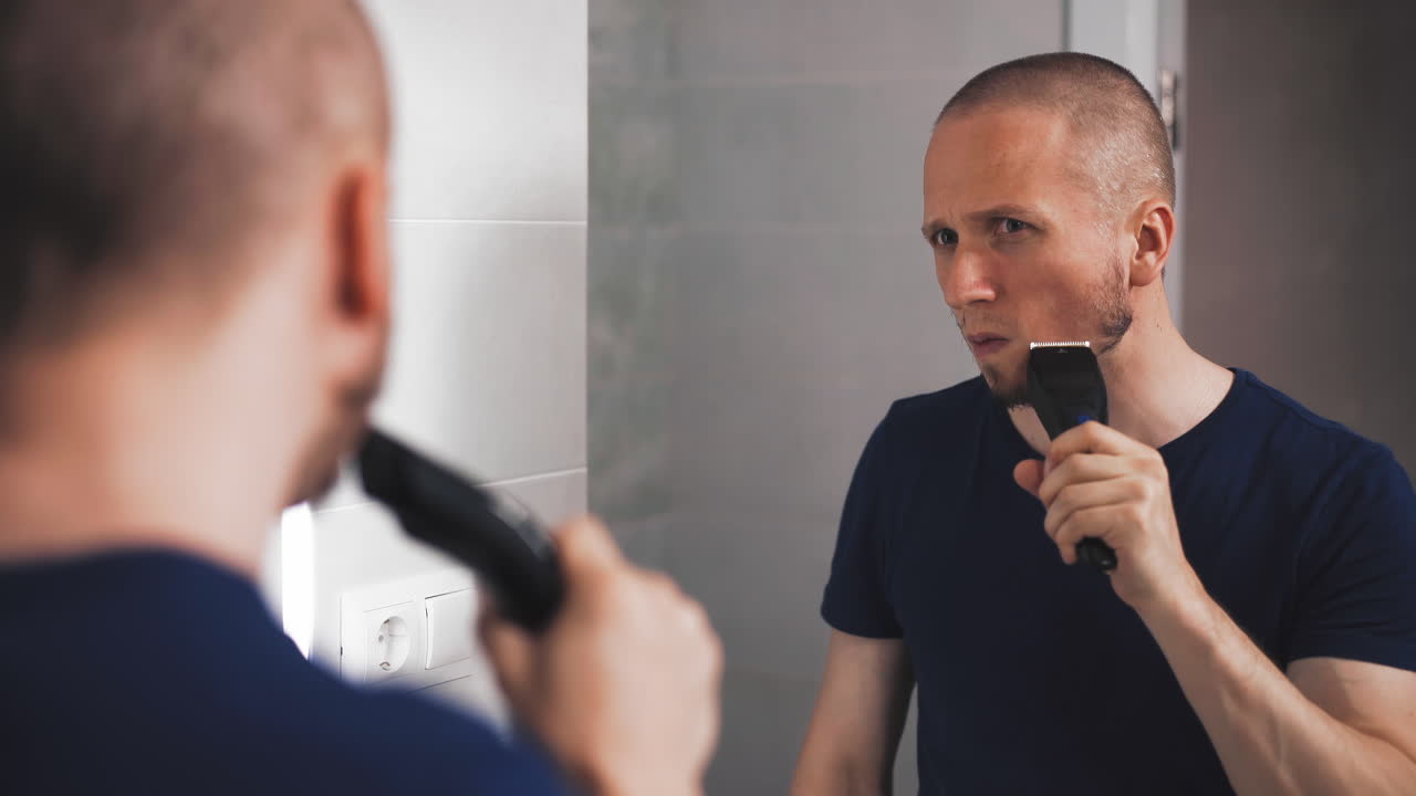 젊은 남자가 큰 거울에서 전기 면도기로 수염을 면도합니다.