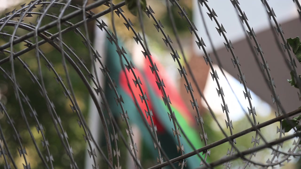 Sharp metal razor wire dominates the foreground as national and political flags wave softly in the background on a Dhaka street. Perfect conceptual b-roll for news stories on regional security.
