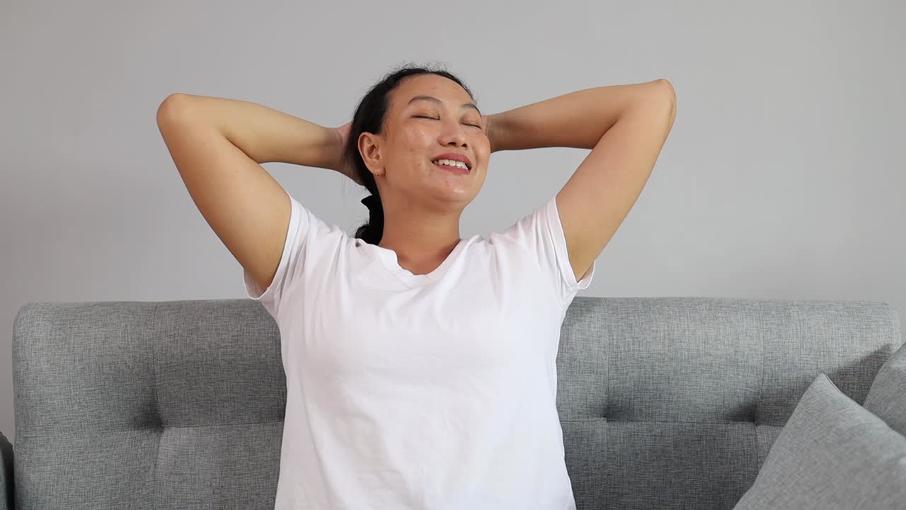 Estilo de vida caseiro relaxante, dona de casa feliz deitada com os braços atrás da cabeça sorrindo. Mulher asiática em poltrona confortável em casa.