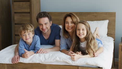 close-up view of cheerful parents and their sons lying on the bed and smiling at camera in the morning