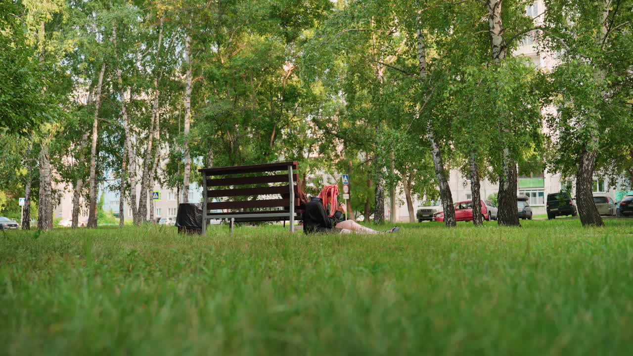 Одинокий пассажир отдыхает возле скамейки в парке с рюкзаком, на переднем плане — широкая лужайка и березы, вдалеке — тихий городской шум, медленная прослеживающая съемка, подчеркивающая тихую паузу, задумчивое настроение и путешествие