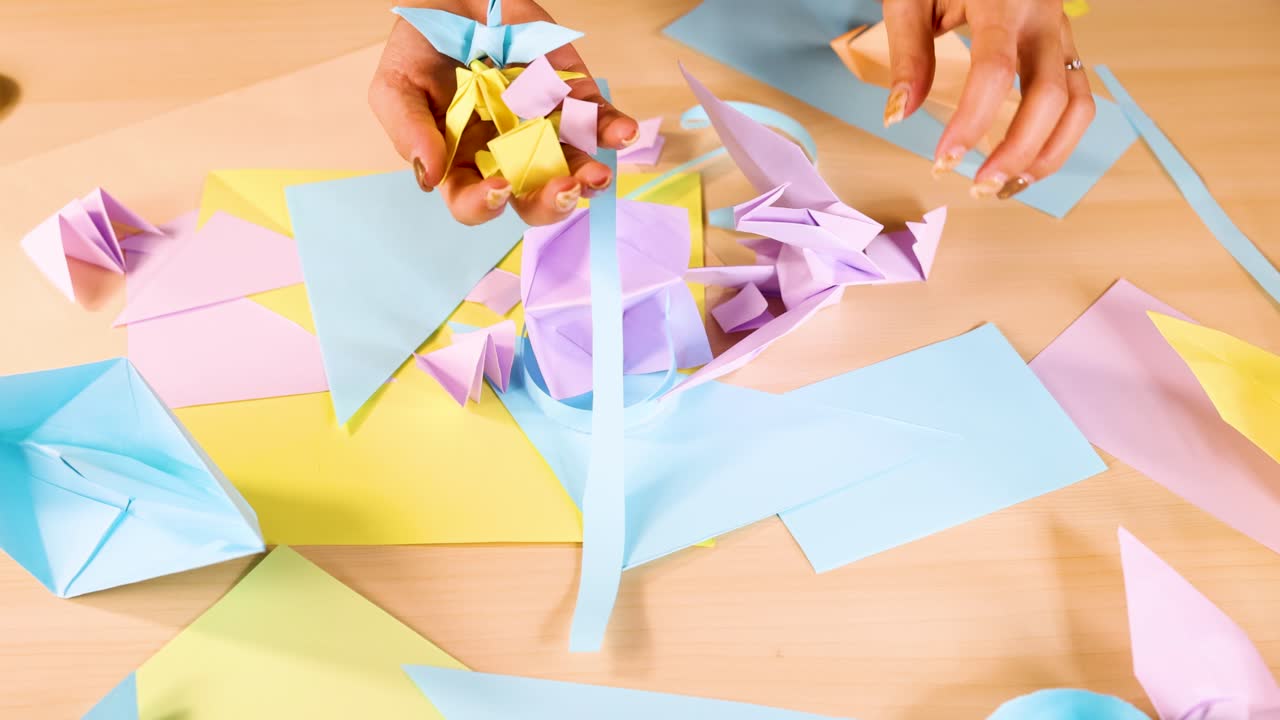 Hands create origami with pastel paper sheets, overhead view, bright lighting, steady camera