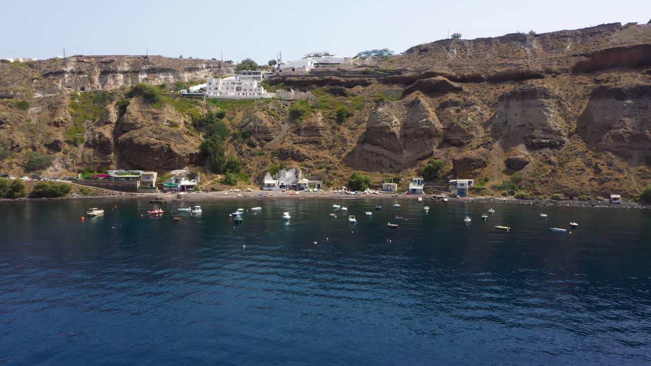 vuelo aéreo hacia atrás que revela la isla griega, acantilado rocoso con villas y barcos de pesca en el mar mediterráneo en la playa de santorini, grecia