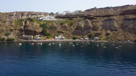 vuelo aéreo hacia atrás que revela la isla griega, acantilado rocoso con villas y barcos de pesca en el mar mediterráneo en la playa de santorini, grecia