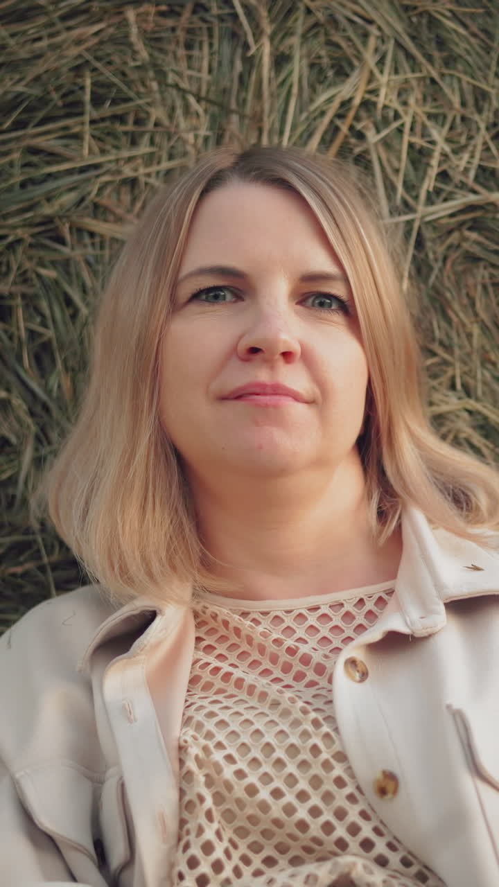 Kvinde i nettop og jakke læner sig op ad en høballe med et varmt smil, nyder et fredfyldt øjeblik på landet, sollyset fremhæver blidt hendes ansigt, hvilket skaber en rolig og afslappet atmosfære.