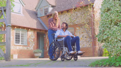 smiling mature couple with man sitting in wheelchair being pushed by woman outside home