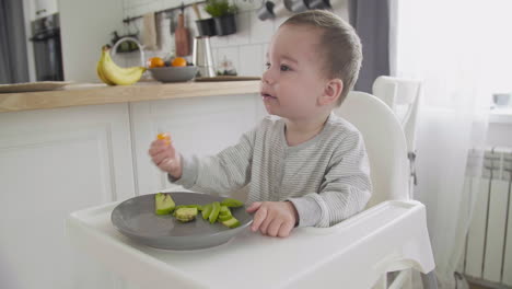 cute baby boy eating clementine and avocado sitting in high chair in the kitchen 1