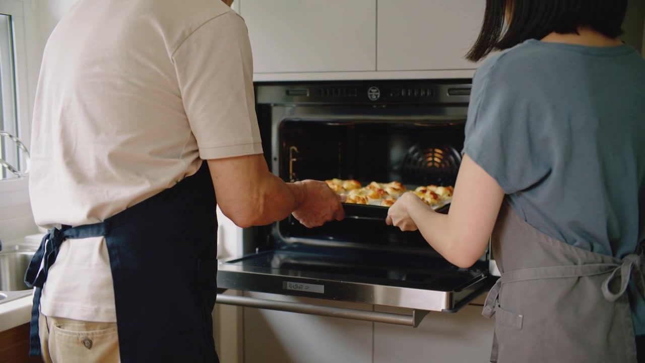 Una coppia con grembiuli toglie una teglia dal forno di casa dopo aver aperto lo sportello per far raffreddare i panini.