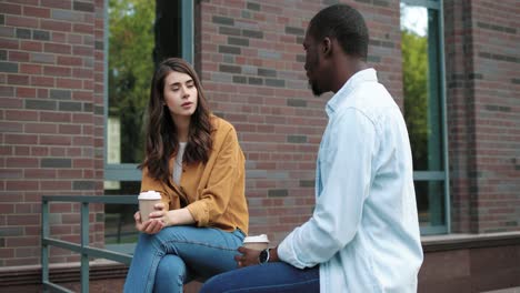 estudiantes caucásicos y afroamericanos hablando y bebiendo café sentados en la calle cerca de la universidad