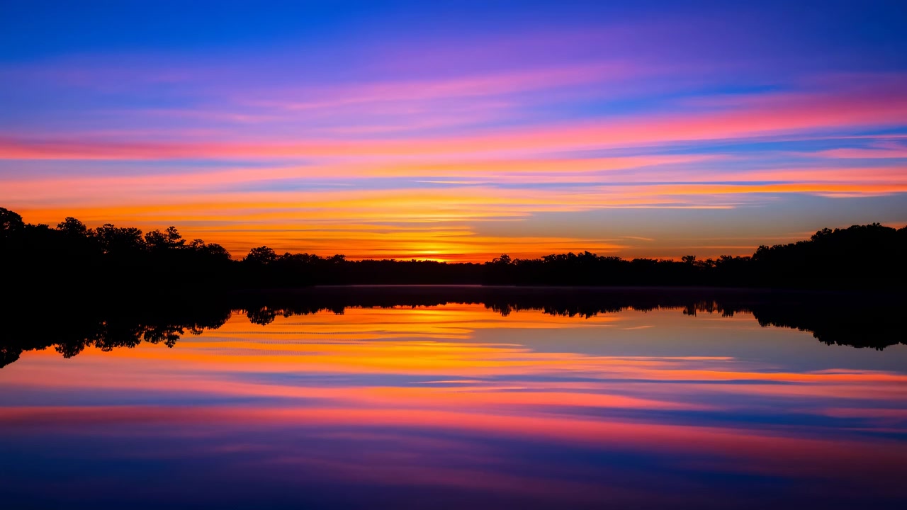 Sunset light spreads across still lake with vivid orange reflection, dark tree line, smooth mirrored water, and soft purple pink sky, creating serene scenic mood for outdoor travel use