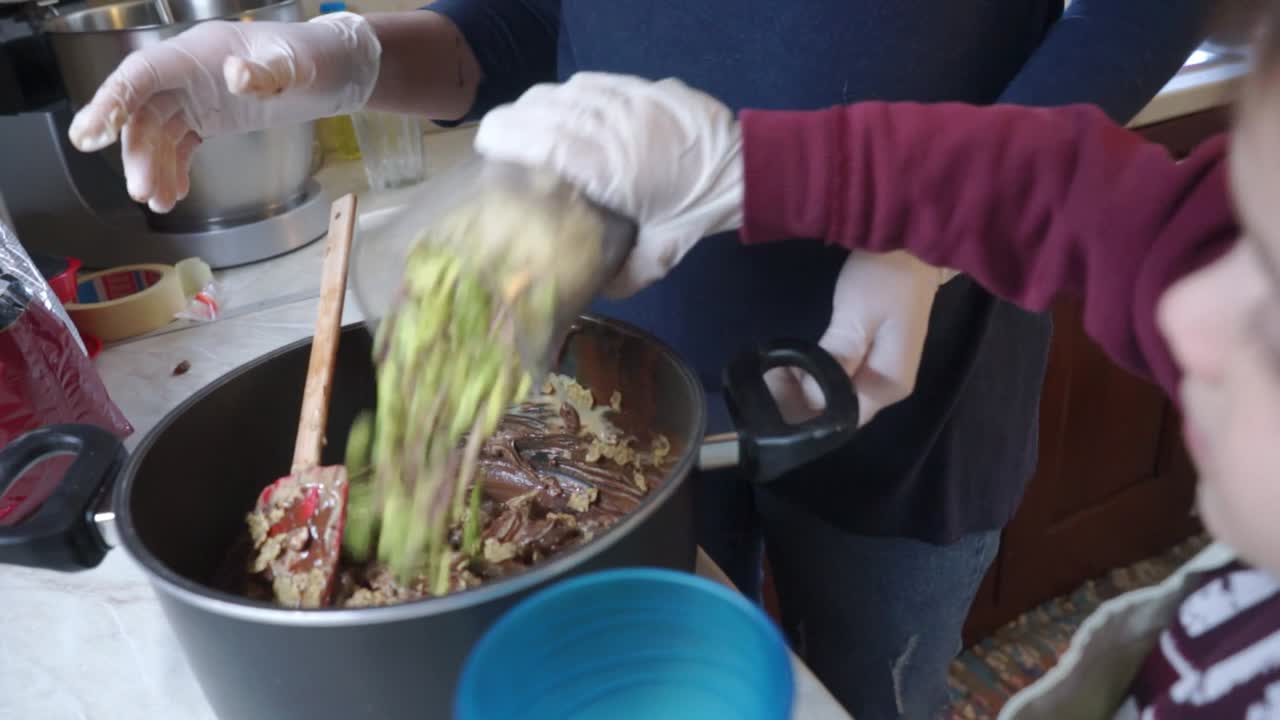 Caucasian toddler boy helping his mother making energy bars in the kitchen pouring ingredients in pot