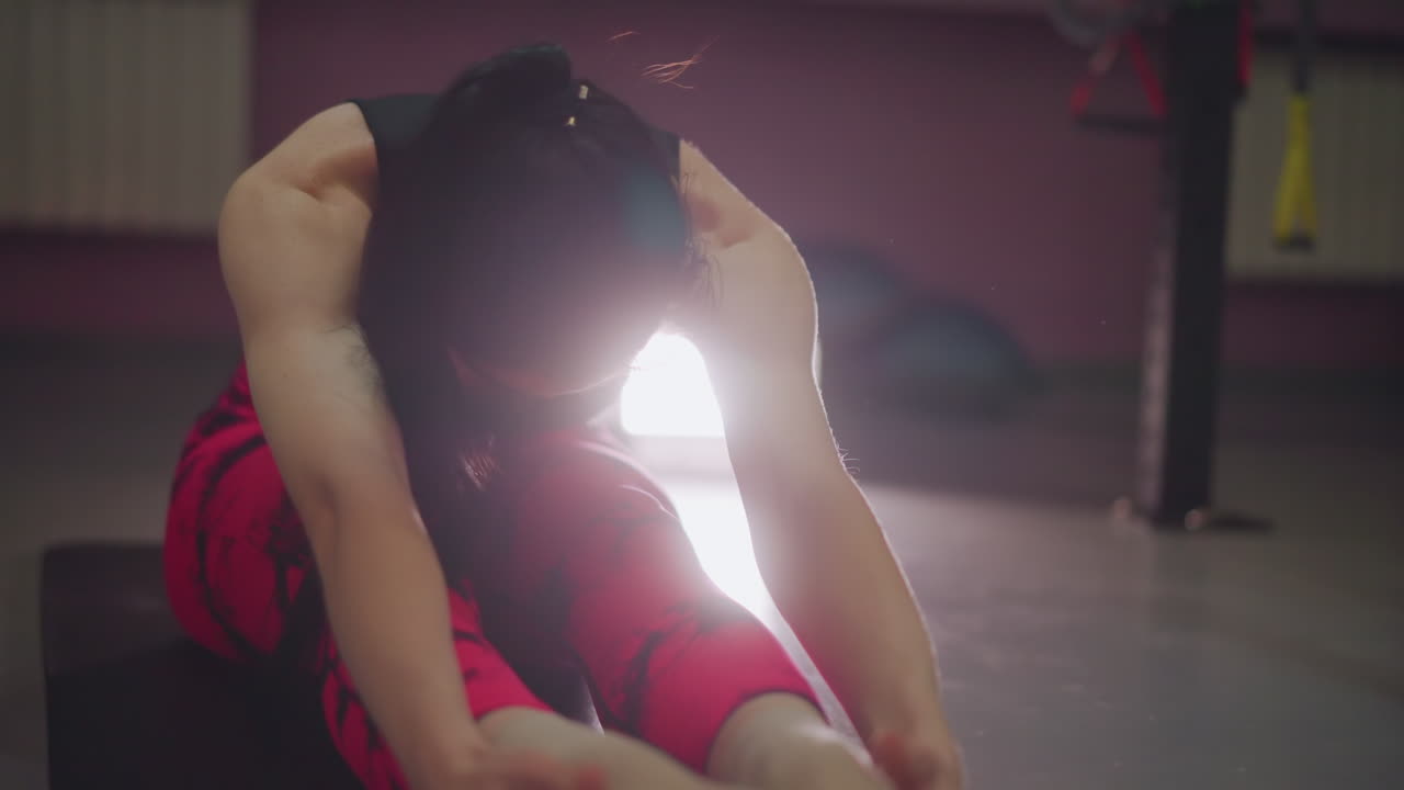 Donna che tocca le dita dei piedi durante lo stretching dei tendini del ginocchio sul tappetino, forte bagliore in controluce ed espressione concentrata, piegamento in avanti profondo per mobilità e progresso, allenamento a piedi nudi in studio con tecnica determinata.