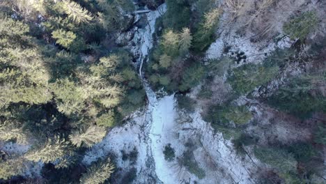 Vista-Aérea-Del-Río-Helado-En-Imponentes-Montañas-Con-Densos-Abetos-Durante-El-Invierno