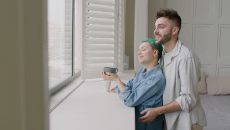 joven hablando y acariciando tiernamente a su novia que sostiene una taza mientras está de pie junto a la ventana del dormitorio