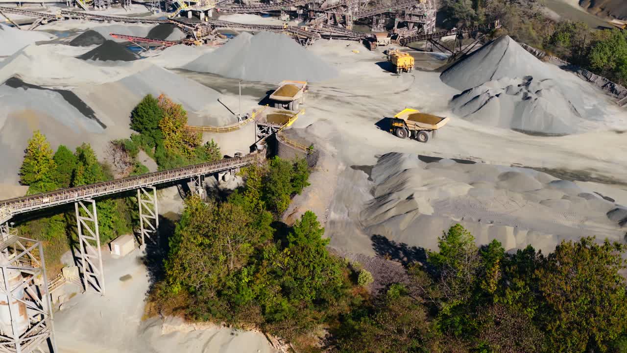 Aerial of quarry processing plant with conveyors and gravel piles, yellow dump truck contrasting, slow motion
