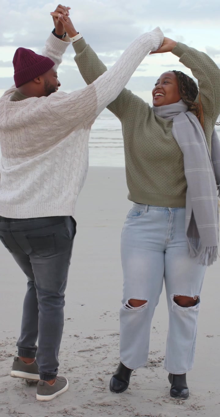 Vertikal video: Afroamerikansk par holder i hånd, snurrer rundt på stranden, legende, mand med hat