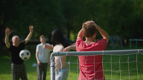 a blurred view of two people in the background while a young boy in red holds his head as a ball crosses over the net, the father playfully runs by, covering his face with his shirt playfully