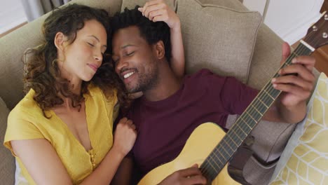 feliz pareja biracial acostada en el sofá juntos y tocando la guitarra