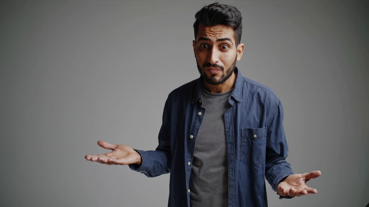 A confused man shrugs in a mid-shot video, wearing casual attire against a plain background
