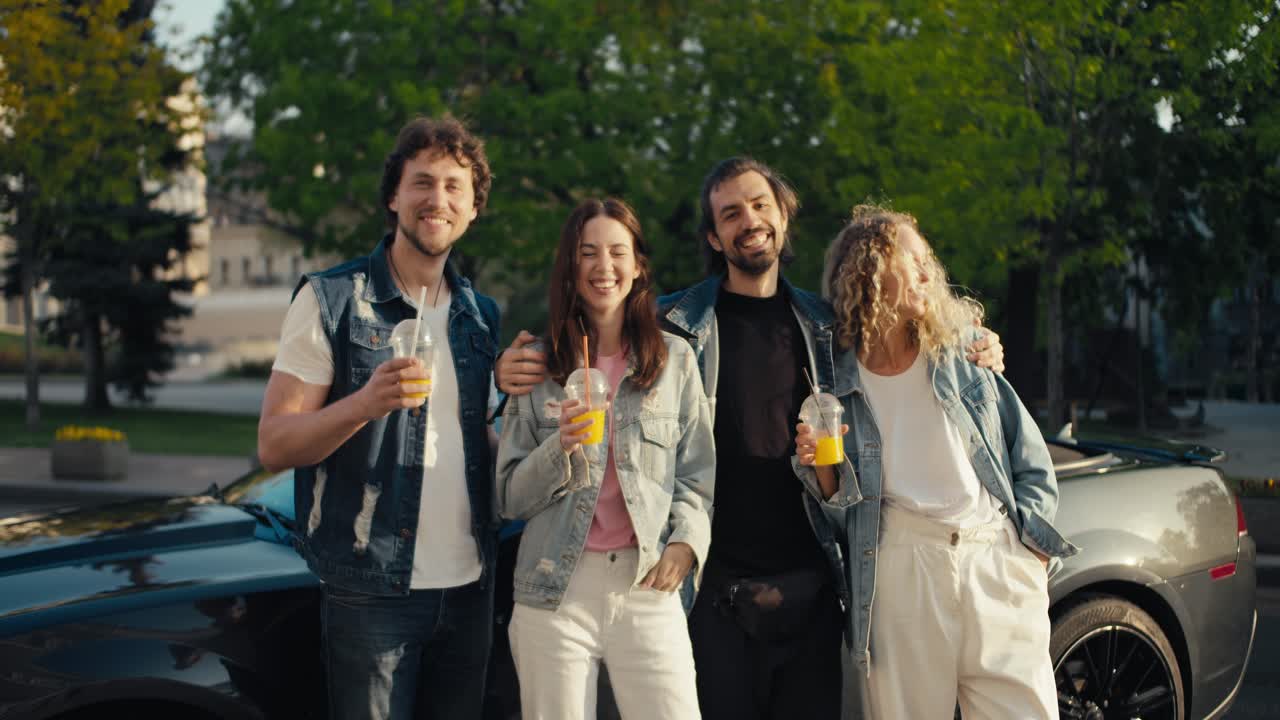 A group of happy friends are standing near their car with yellow cocktails in their hands posing and looking at the camera on a