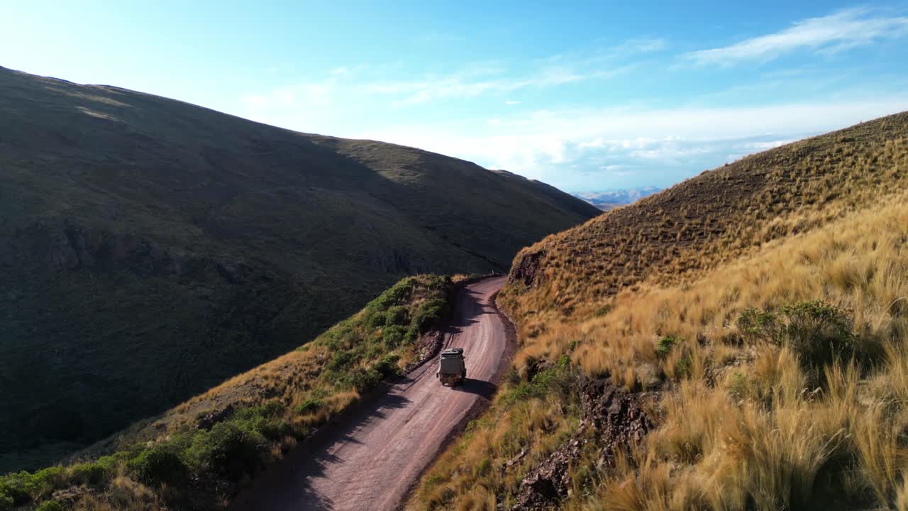 Drone footage following a tuk-tuk on a remote Andean road. The golden hour light highlights the curves of the mountains and the shimmering valley below