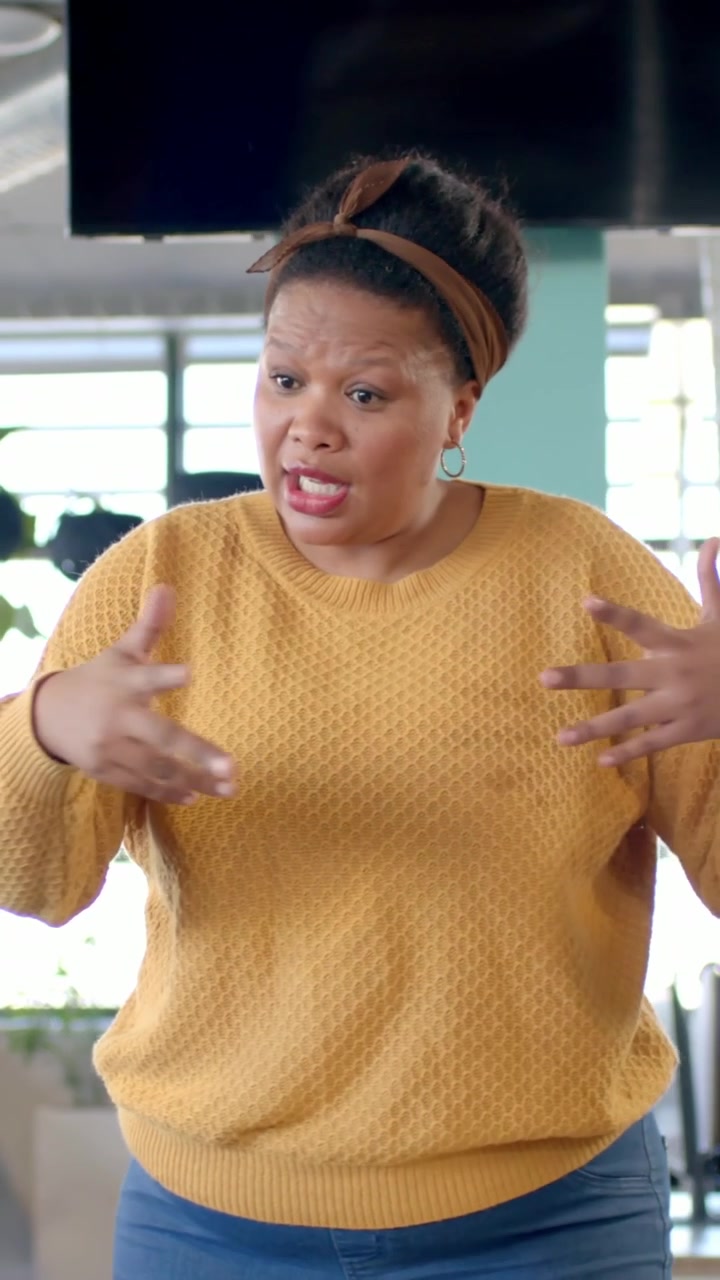 Vertical video: Stepping woman in mustard top leaning on table in office addressing offscreen