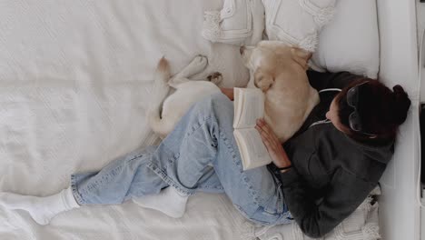 woman relaxing at home, reading a book with her labrador retriever. the dog cuddles with its owner. owner kisses dog's head. a sweet moment of bonding between pet and owner at home. true friendship