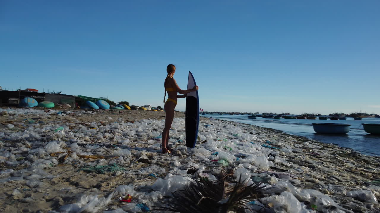 Giovane surfista atletica caucasica in piedi su una spiaggia inquinata da rifiuti di plastica, concetto Madre Terra, inquinamento degli oceani, salva il pianeta