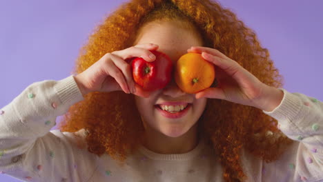 studio portrait of girl holding apple orange in front of eyes against purple background