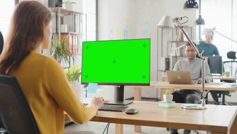 over the shoulder: creative young woman sitting at her desk using desktop computer with mock-up green screen. in the background bright office where diverse team of young professionals work on computers