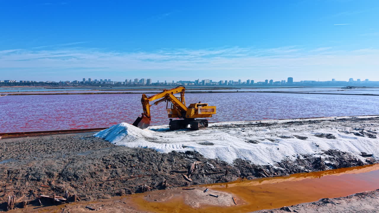 Escavatore giallo al lavoro su un cumulo di sale bianco in una salina. Un escavatore industriale scava sale bianco su un argine tra vasche di evaporazione rosa sotto un cielo azzurro limpido.