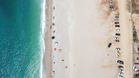 cars parked close to the beach with people relaxing on the shore and playing with the waves