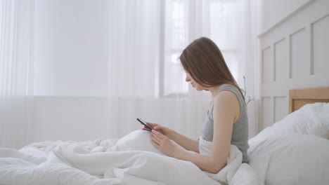 Young-caucasian-woman-sitting-on-the-bed-in-the-morning-feels-bad.-A-young-woman-is-making-a-smartphone-call-to-her-doctor.