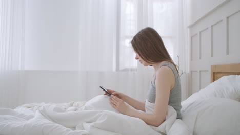 young caucasian woman sitting on the bed in the morning feels bad. a young woman is making a smartphone call to her doctor.