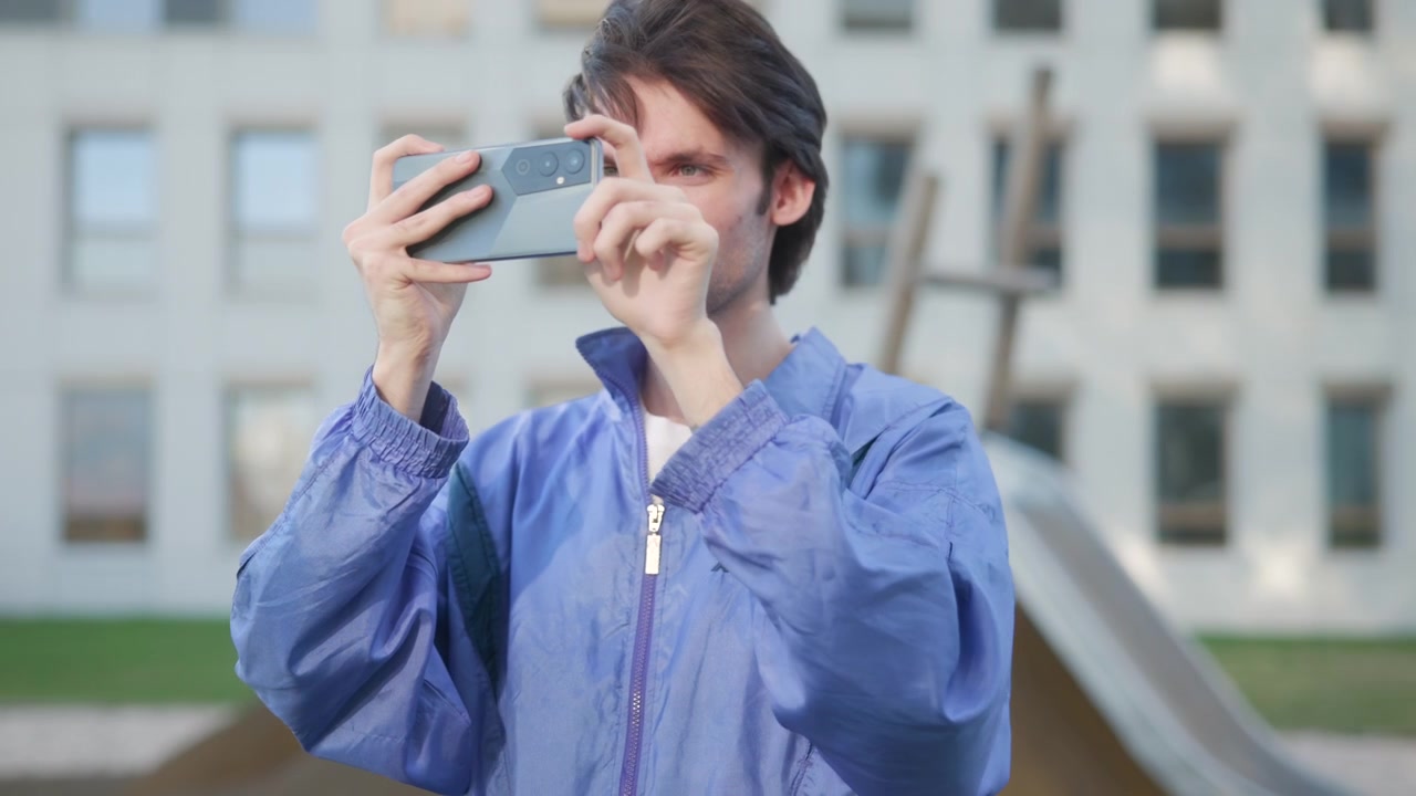 Homem adulto de jaqueta azul está em pé ao ar livre segurando um smartphone horizontalmente, parecendo tirar uma foto ou gravar em um ambiente urbano contemporâneo.
