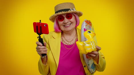 una mujer mayor con una chaqueta amarilla y cabello rosa sonríe para una selfie con un flotador de pato y un flotador del anillo