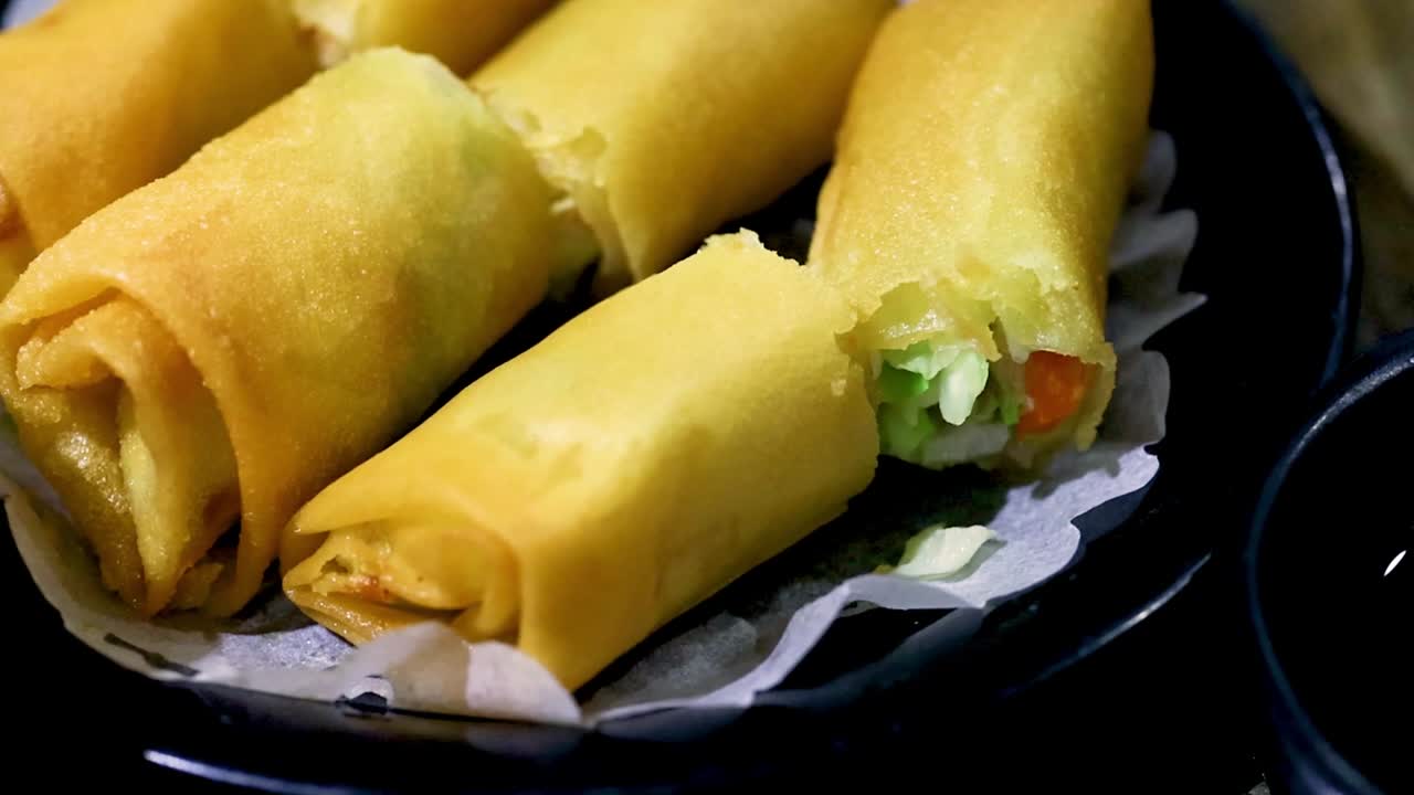 A detailed view of golden, crispy spring rolls served on a black plate with a paper liner.
