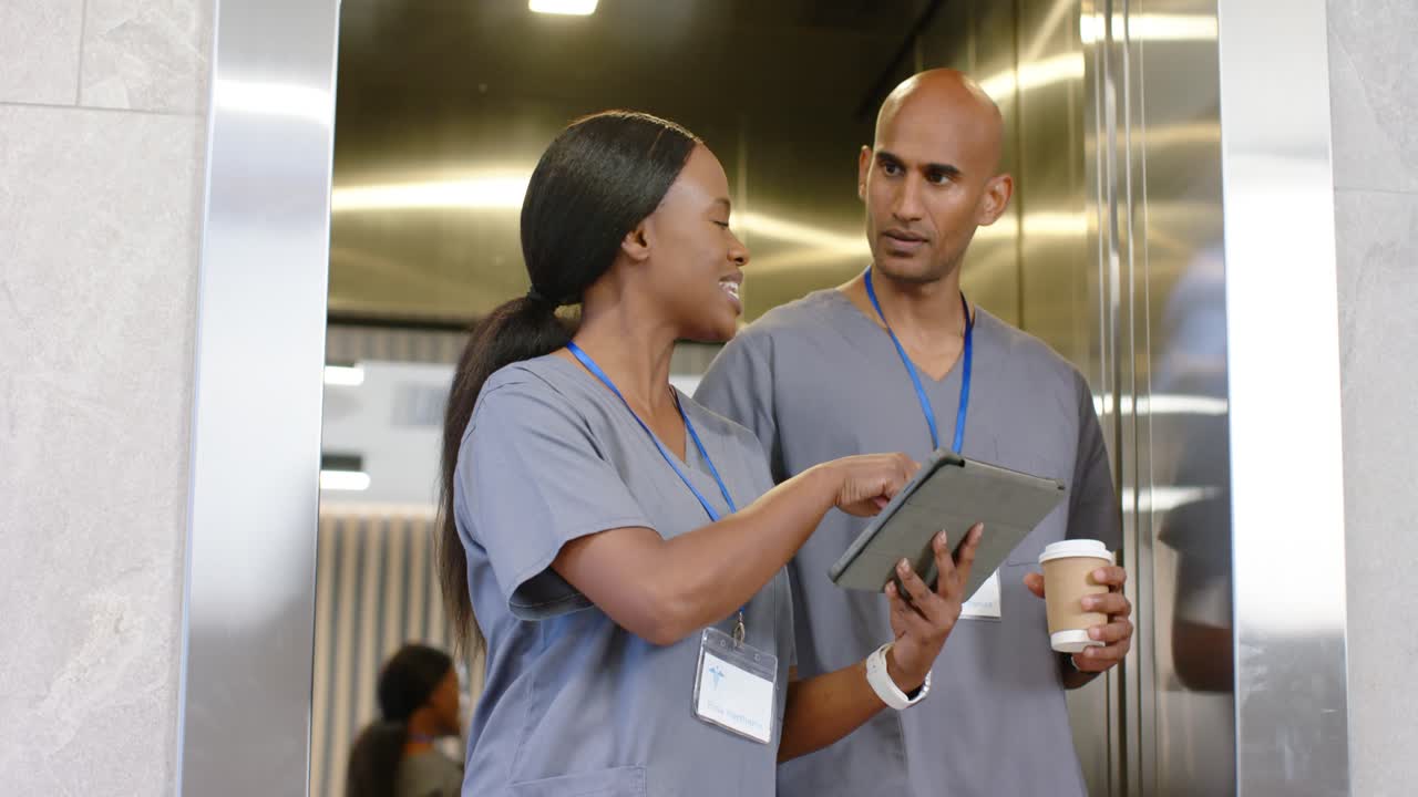 Elevator opening, two staff in scrubs, woman tapping tablet and man holding coffee, discussing care