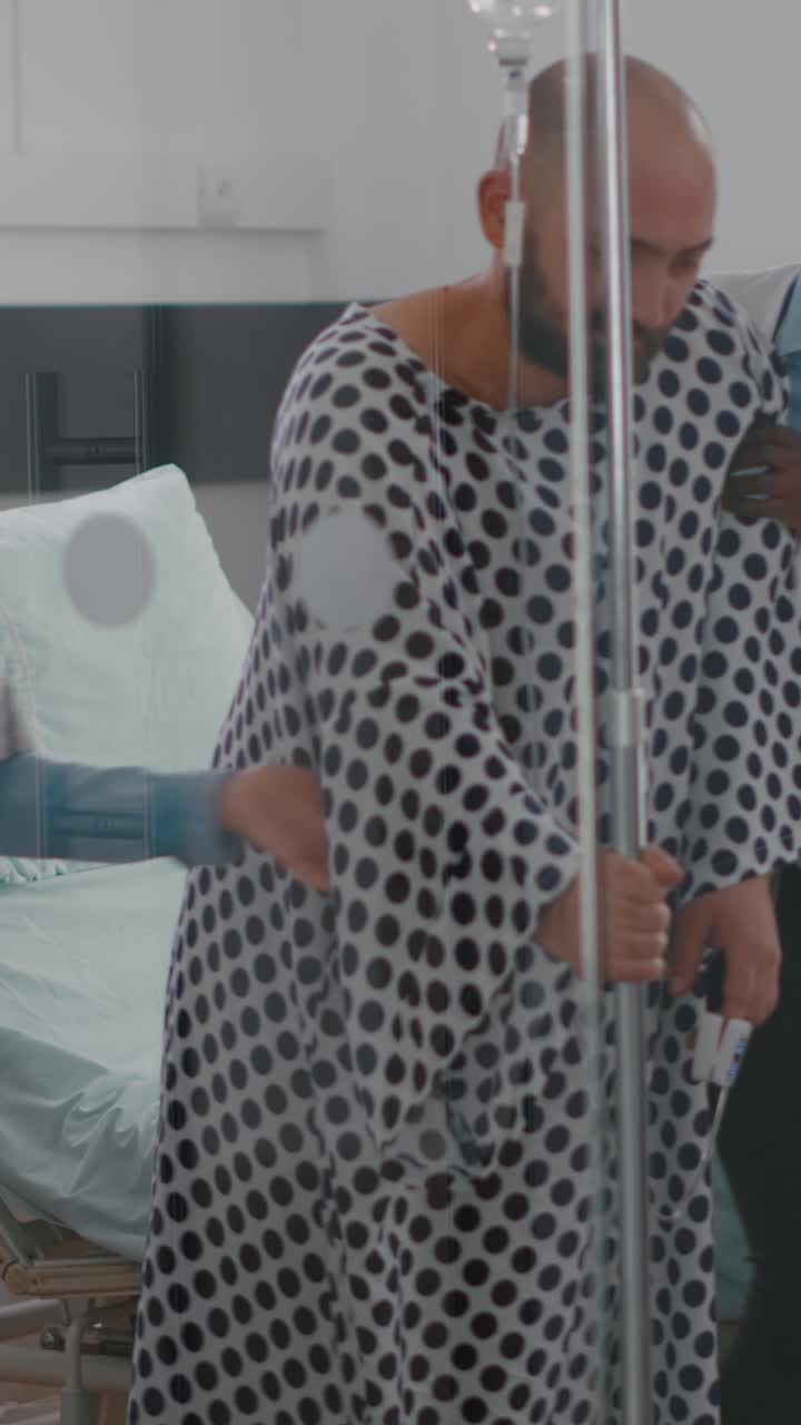 Vertical video: Practitioner doctors helping patient to stand up from bed holding intravenous IV fluid drip bag