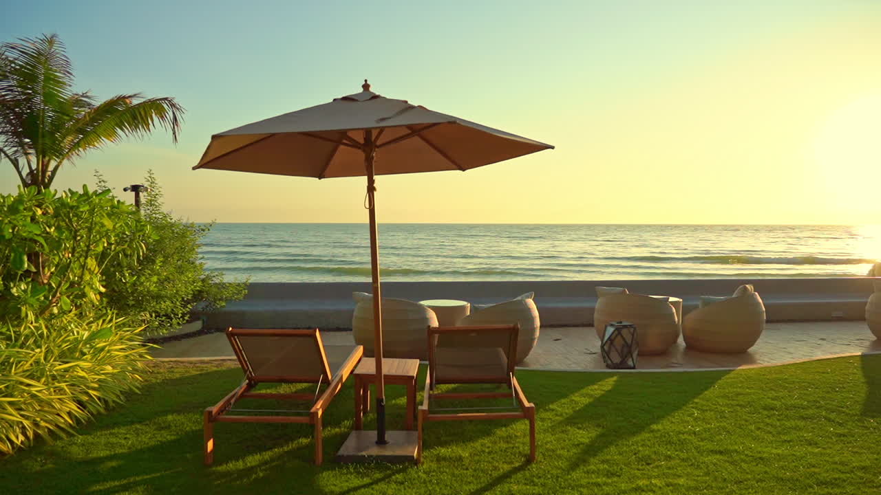 Verde erba lussureggiante e paesaggio tropicale di un bellissimo resort su un'isola con sedie che si affacciano sulla riva dell'oceano al tramonto, con la luce dorata che risplende sulla superficie del mare