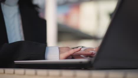 close-up of lady's hands with polished nails and delicate rings typing on laptop keyboard outdoors, placed on wooden table with blurred background