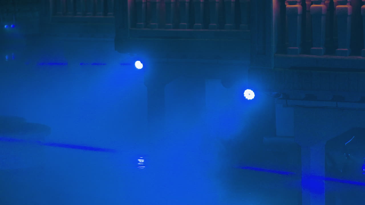 Kabut panggung biru dan lampu sorot menciptakan suasana klub sinematik yang muram, lengkap dengan balkon dan pagar industrial. Sorotan cahaya berkabut serta berkas laser menembus asap tebal, diiringi penerangan menyebar dan nuansa kobalt.