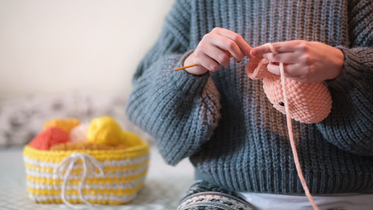 Woman in gray sweatshirt is knitting a toy using hooks and pink yarn
