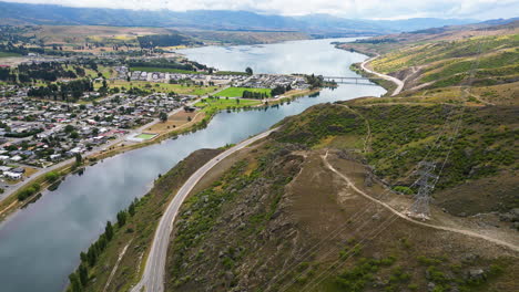 Aerial-droone-backward-moving-shot-over-Cromwell-town-alongside-Dunstan-lake-in-New-Zealand-with-the-view-of-a-winding-road-passing-along-the-hilly-terrain