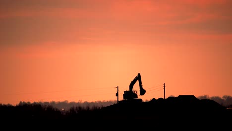 Excavator-scooping-and-dumping-earth-soil-against-a-background-of-orange-sky