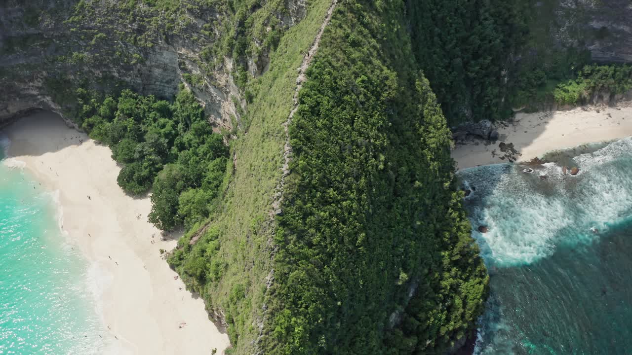 Aerial drone shot in the iconic Kelingking Beach, the 'T-Rex' shaped headland, and the towering limestone cliffs of Nusa Penida. Turquoise Indian Ocean crashes against the rocks and white-sand beach